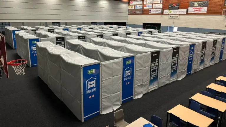 Rijen grijze slaapcabines met blauwe deuren (BED-STAY) opgesteld in een sporthal