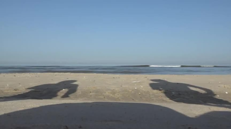 Strand met rustige zee en blauwe lucht, schaduwen van twee mensen op het zand zichtbaar.