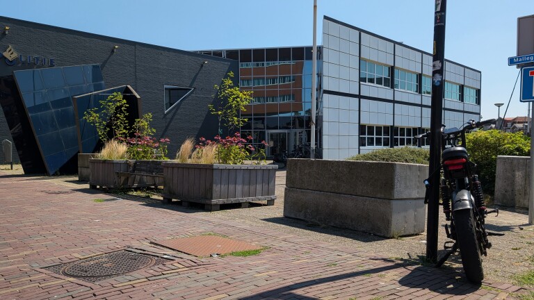 Modern gebouw met schuine glazen gevel en witte panelen, met bloembakken op het plein en een geparkeerde motorfiets