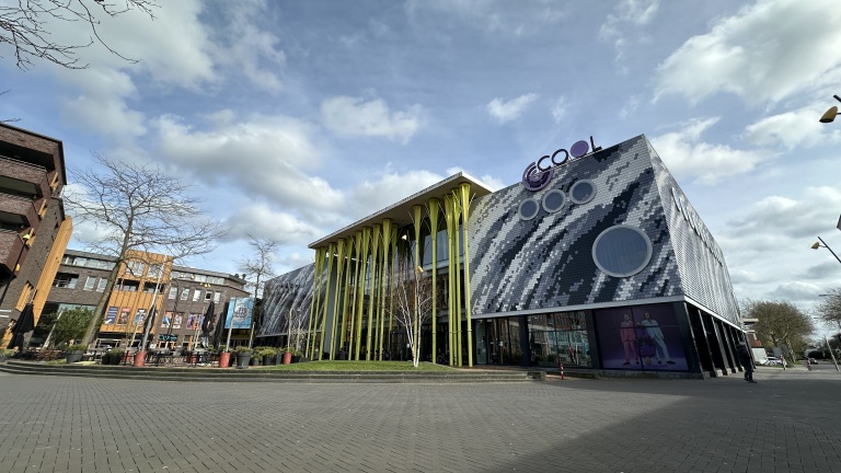 Modern gebouw met opvallende gele zuilen en een mozaïekachtige gevel in een stedelijke omgeving onder een blauwe lucht.
