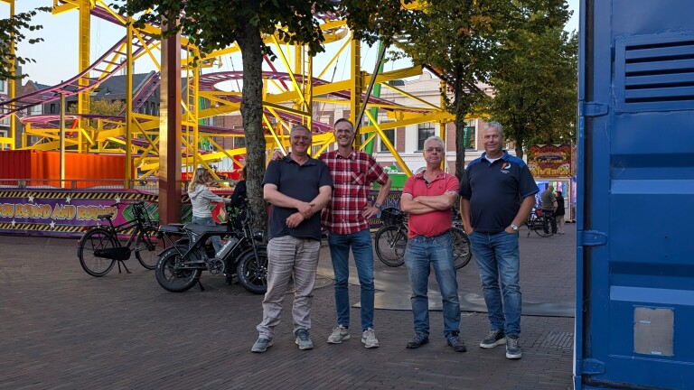 Vier mannen poseren op een plein voor een kleurrijke kermisattractie met gele constructie en paarse rails