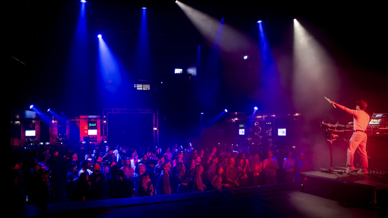 DJ op podium wijst naar het publiek in een donkere clubzaal met blauwe en paarse spotlights en een volle dansvloer