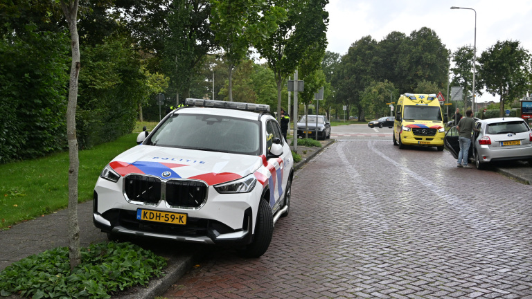 Politieauto en ambulance op een straat, met enkele mensen bij een stilstaande auto langs de weg