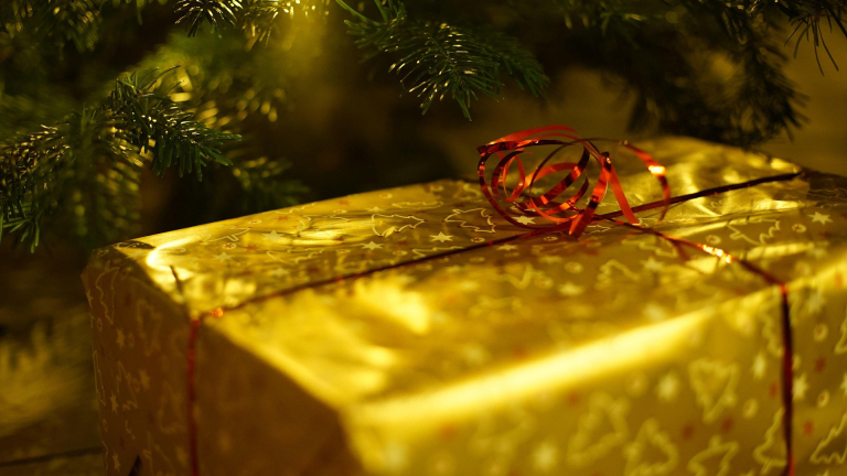Goudkleurig kerstcadeau met rood krullint onder de takken van een kerstboom in warm licht