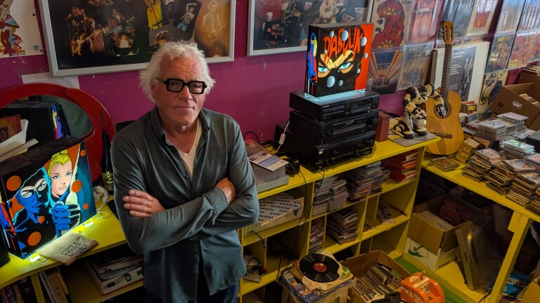 Man met grijs haar en zwarte bril in muziekruimte vol cd’s, platen, posters, stereo-installatie en gitaar