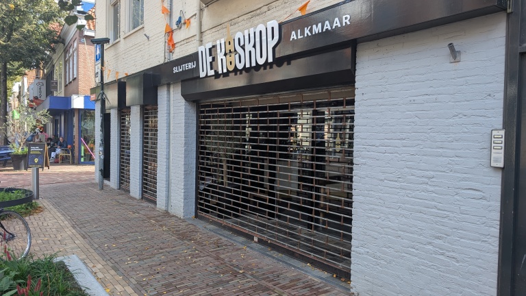 Gesloten winkelpui met rolluik en gevelbord “Slijterij De Haskop Alkmaar” aan een straat met stoep en bomen