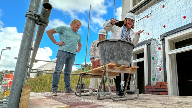 Drie mensen met bouwhelmen op een steiger bij een huis in aanbouw, met speciekuip op een kruiwagen