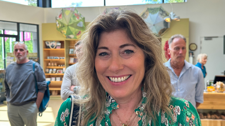 Lachende vrouw met golvend haar in groene blouse, binnen in een winkel of café met mensen en schappen op de achtergrond