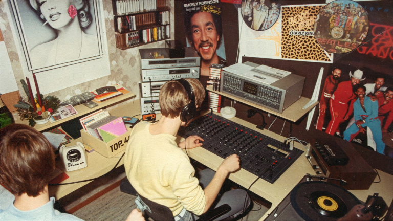 Jongen met koptelefoon bedient een mengpaneel in een kamer vol lp’s, cassettes en muziekposters