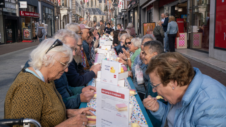 Lange tafel in winkelstraat met veel mensen die samen gebak eten; bordje met tekst Wie jarig is trakteert