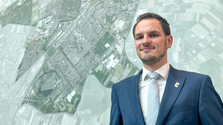 Man in blauw pak met stropdas en reversspeld, staand voor een grote luchtfoto- of stadskaart