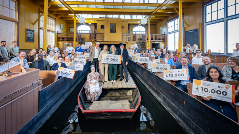 Groepsfoto in houten veilingzaal; deelnemers houden cheques van €10.000 vast, centraal een cheque van €21.000