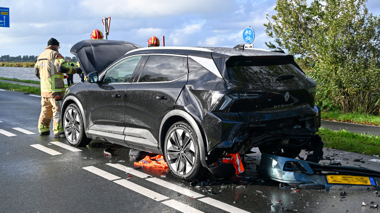 Beschadigde zwarte auto na aanrijding op natte weg, met brandweer en brokstukken achter de auto
