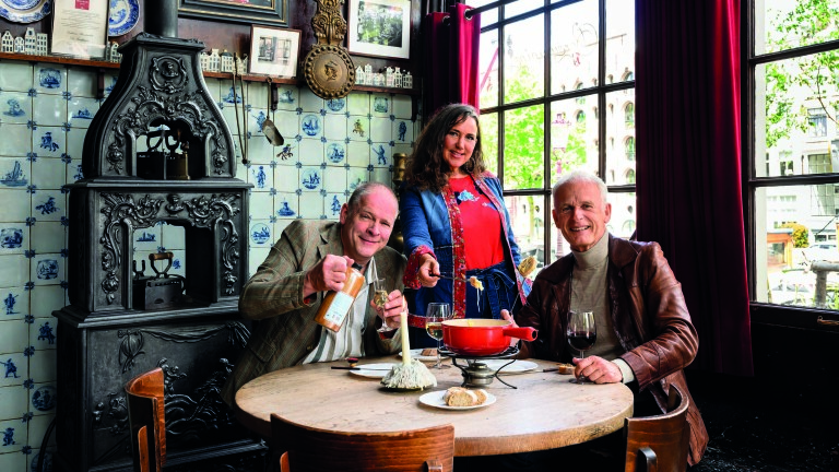 Drie mensen aan tafel in een gezellig restaurant met fonduepan, wijn en oud-Hollandse tegels op de muur