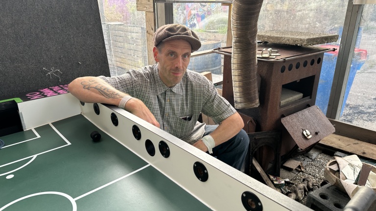 Man met pet leunt op een tafelvoetbaltafel in een rommelige werkruimte met een houtkachel op de achtergrond