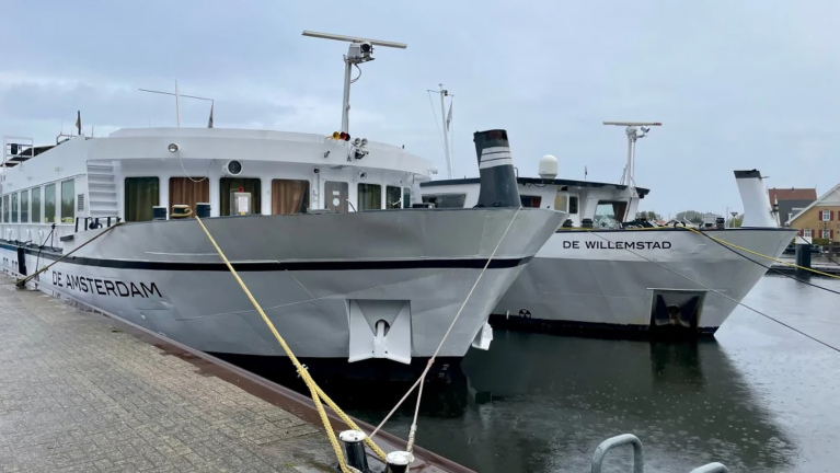 Twee grijze schepen, De Amsterdam en De Willemstad, liggen aangemeerd in een haven op een bewolkte dag