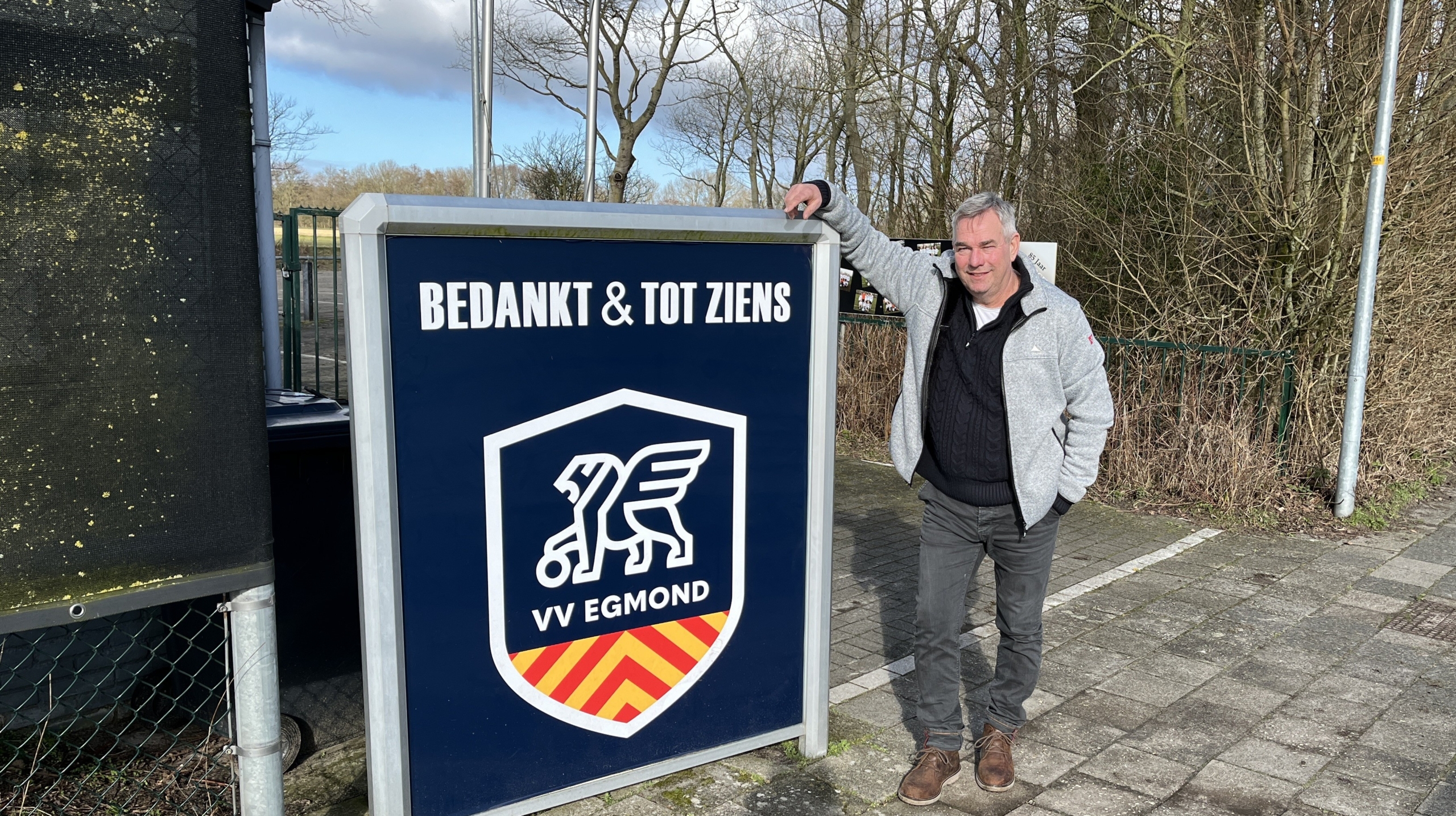 Man bij een bord met de tekst "Bedankt & Tot Ziens" en het logo van VV Egmond, op een buitenlocatie met bomen.