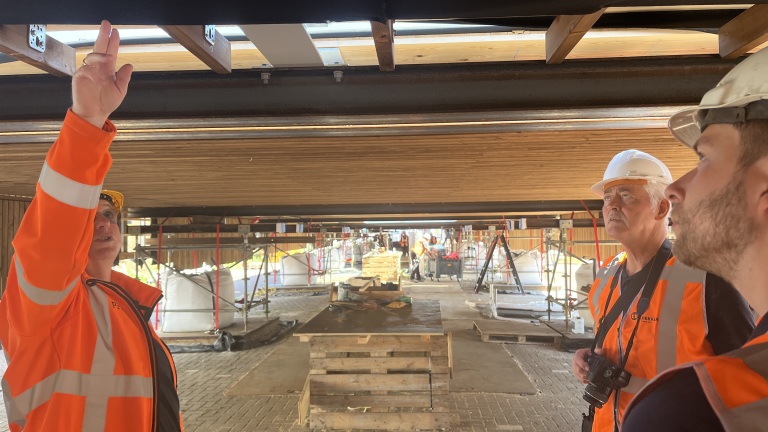 Drie mannen in oranje veiligheidskleding en helmen gebogen onder een constructie op een bouwplaats, waarbij één naar boven wijst.