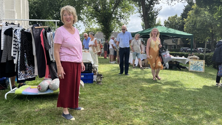 Een buitenmarkt op gras met een vrouw in een roze geruite blouse en rode broek die bij een kledingrek staat, omringd door mensen bij verschillende kraampjes.