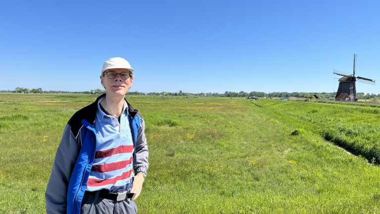 Man met een pet en bril staat in een groen weiland onder een heldere blauwe lucht, met een traditionele houten windmolen op de achtergrond.