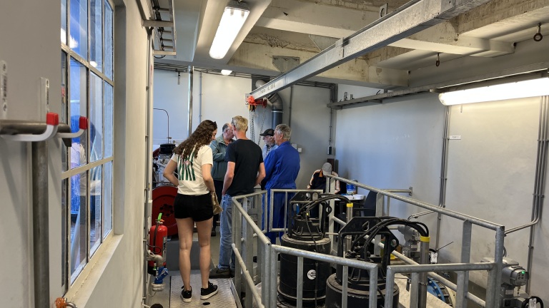 Groep mensen in een technische ruimte met metalen apparatuur en buizen, verlicht door kunstlicht.