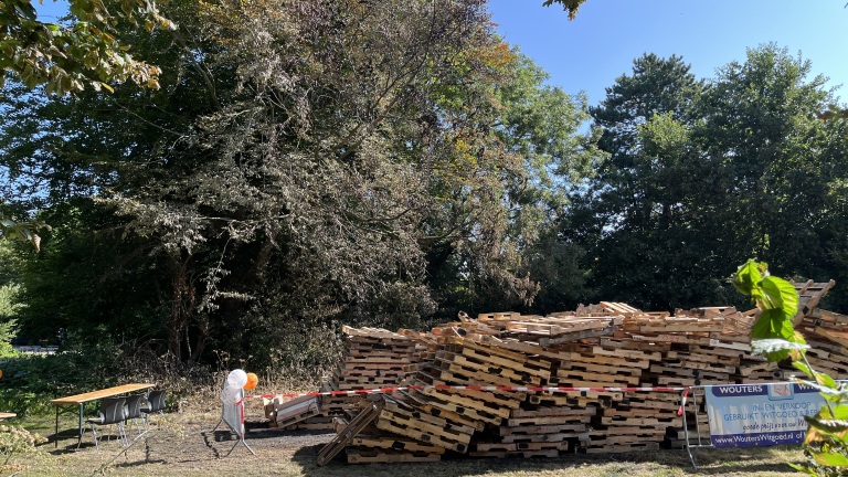 Een grote stapel houten pallets staat op een grasveld naast bomen, met een reclamebord en een tafel met stoelen in de buurt.