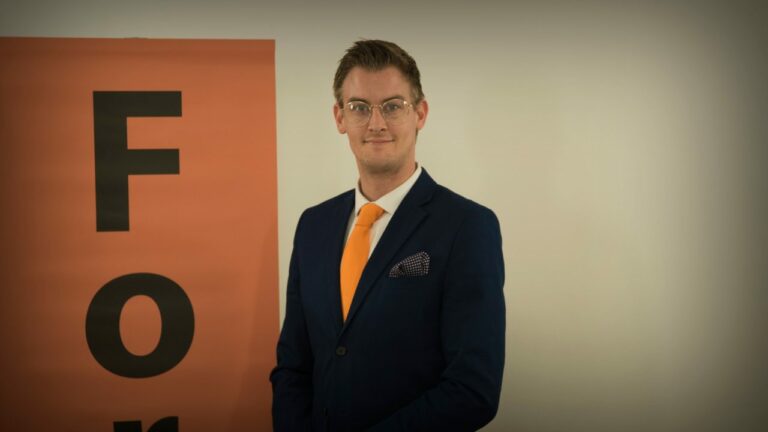 Een man in een donkerblauw kostuum met een fel oranje stropdas staat naast een oranje bord met de letters 'F' en 'O'.