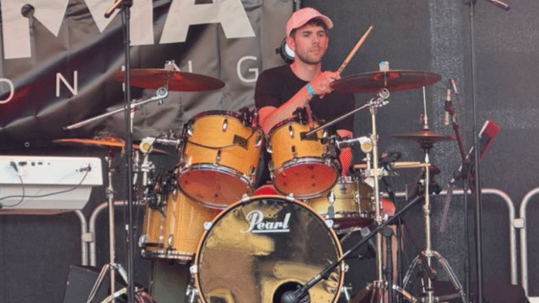 Een drummer met een roze pet speelt op een Pearl drumstel tijdens een live optreden op een podium.
