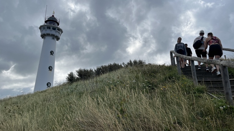 Lighthouse on a cloudy day beside a grassy hill with a group of people descending wooden stairs.