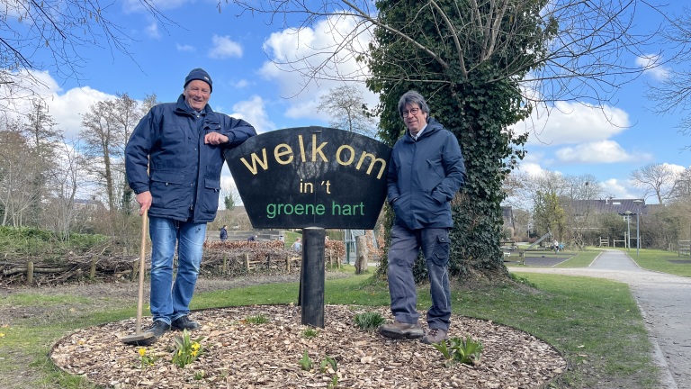 Twee mannen poseren naast een bord met de tekst "welkom in 't groene hart" in een parkachtig gebied.