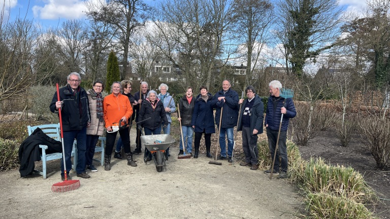 Groep mensen met tuingereedschap in een tuin, omringd door bomen en droge planten.