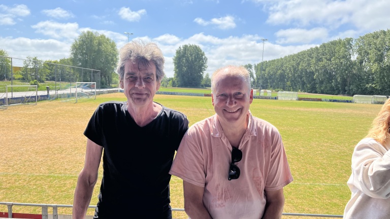 Twee mannen staan naast elkaar op een zonnig sportveld, met een heldere blauwe lucht en groene bomen op de achtergrond, en dragen respectievelijk een zwart en een lichtroze shirt.