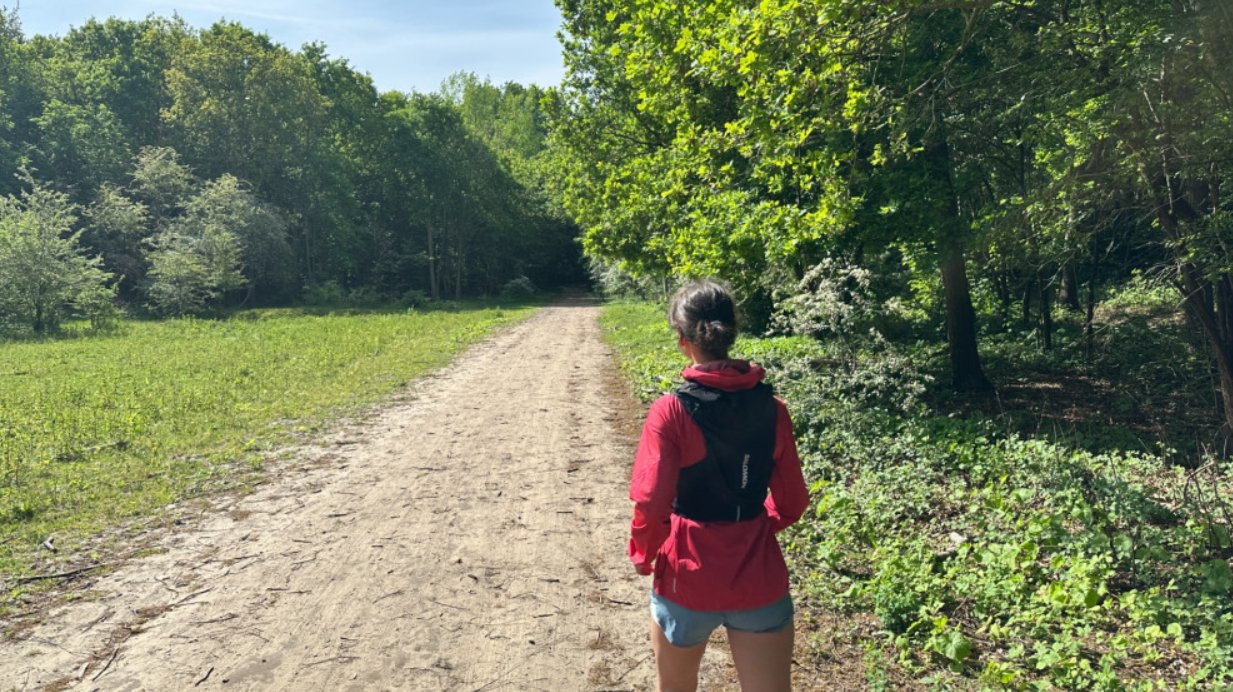 Vrouw gekleed in een rode jas en korte broek loopt op een zandpad door een groene bosrijke omgeving onder een heldere hemel.