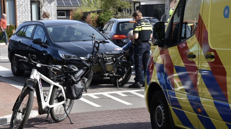Een politieagent onderzoekt een ongeval tussen een zwarte auto en een fiets. Een ambulance met rood en blauw gestreept patroon staat dichtbij. Twee fietsen staan geparkeerd naast de auto.