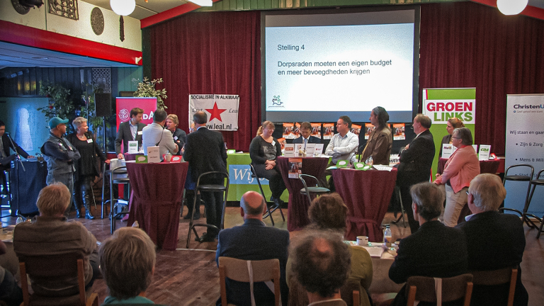 Gezelschap mensen bij een politieke discussie met banners van verschillende Nederlandse partijen in een zaal.