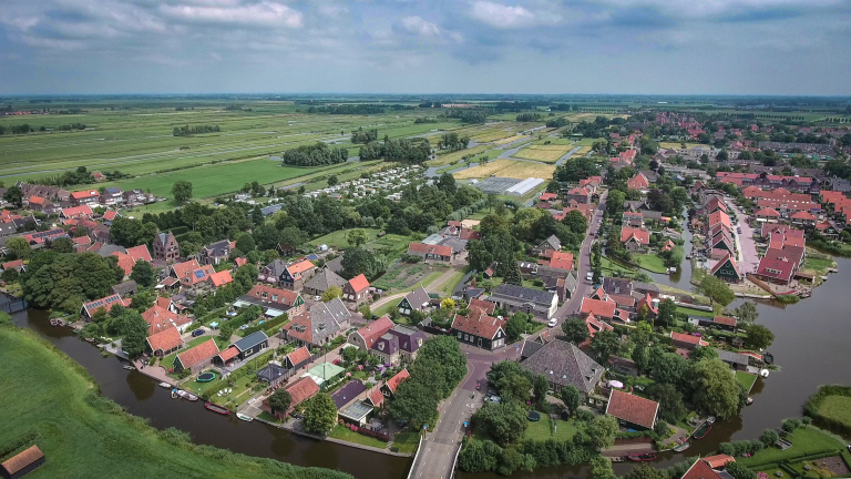 Luchtfoto van een Nederlands dorp met rode daken, grachten en groene weilanden op de achtergrond.