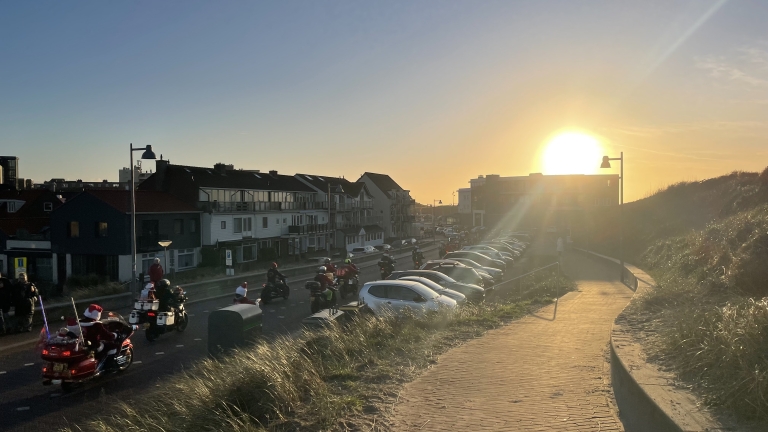Motorrijders in kerstmannenpakken rijden langs een straat bij zonsondergang, met geparkeerde auto's aan de rechterkant en huizen aan de linkerkant.