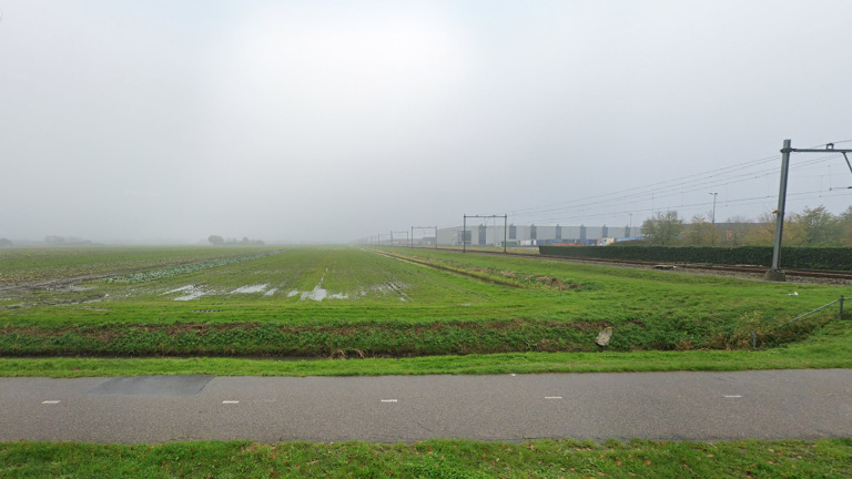 Een mistig landschap met groene velden, een weg op de voorgrond en een spoorlijn met masten langs de zijkant. Op de achtergrond zijn een aantal gebouwen zichtbaar.