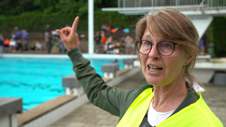 Een vrouw met bril en geel veiligheidsvest wijst met haar vinger naar iets in de verte bij een buitenzwembad, met mensen op de achtergrond.