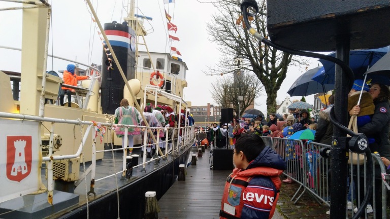 Aankomst in Sinterklaas met mensen op een pier en Pieten op een boot; regenachtige dag met parasols.