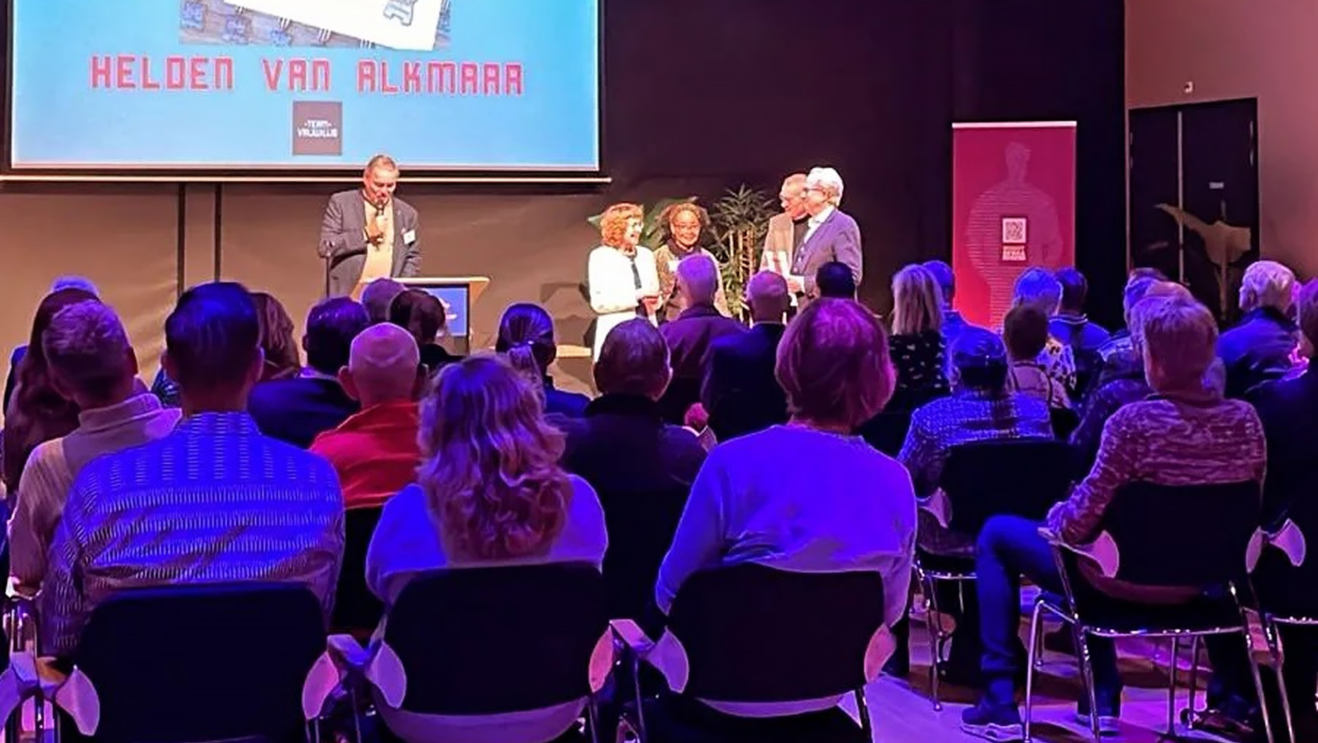 Mensen zitten in een zaal tijdens een evenement met een scherm waarop "Helden van Alkmaar" staat.
