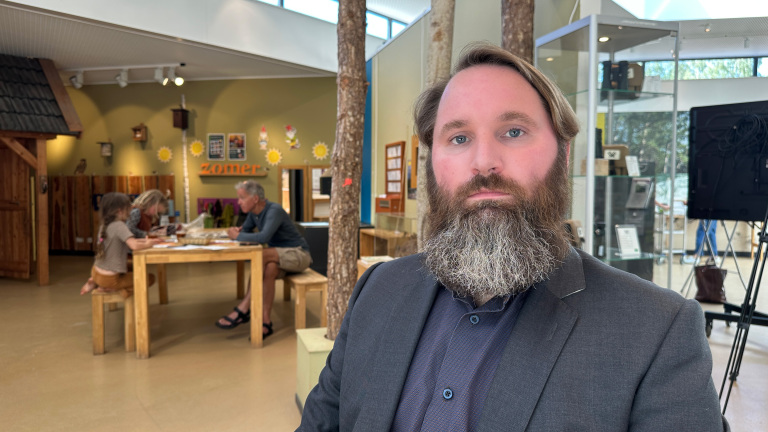 Man met baard in colbert in een museumruimte; op de achtergrond zitten bezoekers aan een tafel en hangt tekst “zomer”