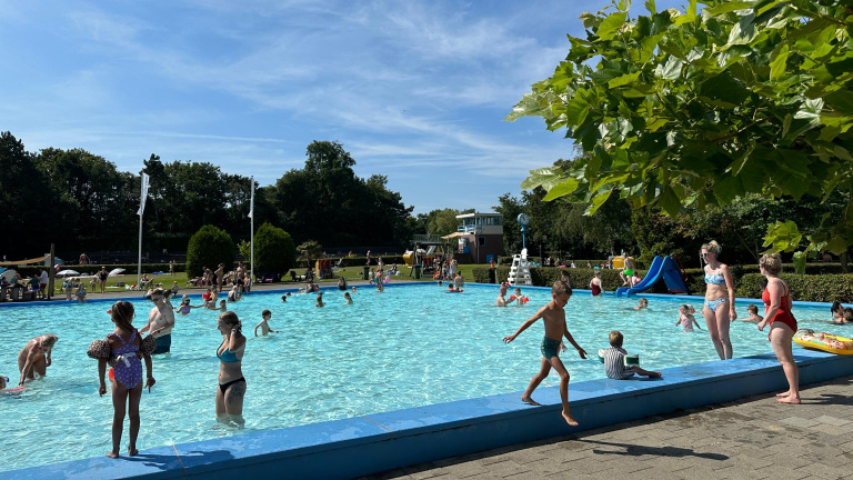 Kinderen en volwassenen spelen in een buitenzwembad op een zonnige dag, met groen rondom en een blauwe glijbaan zichtbaar.