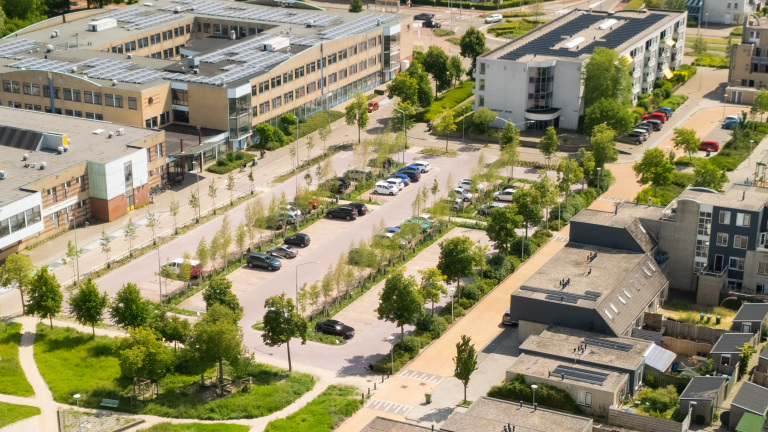 Parkeerplaats met auto's naast een groot gebouw met zonnepanelen op het dak, omringd door groen en andere gebouwen.