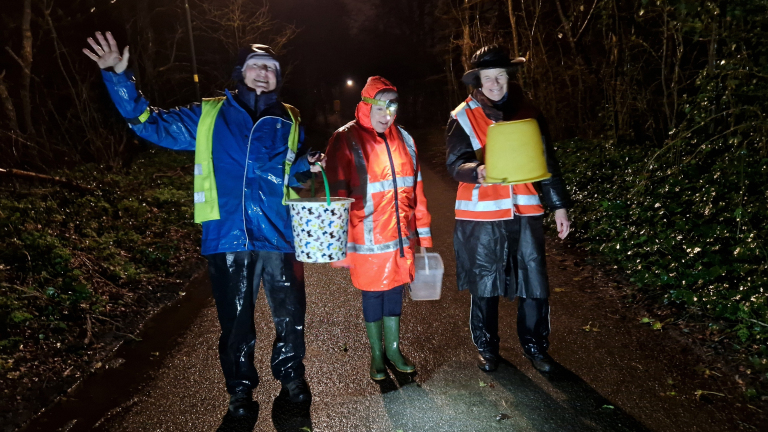 Drie mensen in regenjassen en reflecterende vesten staan 's nachts op een natte weg met emmers.