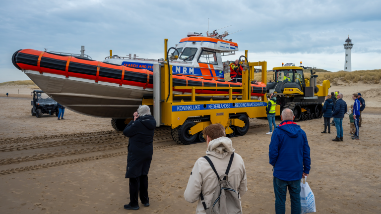 Reddingsboot op een strand met toeschouwers, sleepvoertuig en vuurtoren op de achtergrond.