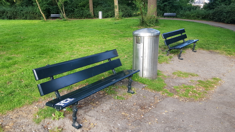 Twee parkbanken en een prullenbak op een groen grasveld met bomen en struiken.