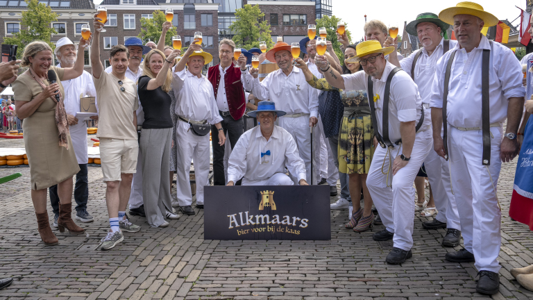 Groep mensen met bierglazen omhoog tijdens het Alkmaars kaasfeest, inclusief kaaskeurders in witte kleding en gekleurde hoeden, en een bord met de tekst "Alkmaars bier voor bij de kaas."