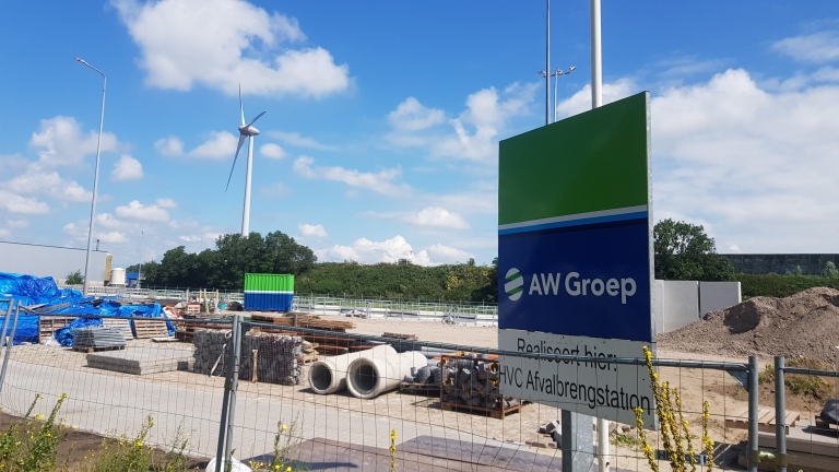 Bouwplaats met bord van AW Groep en windturbine op de achtergrond.