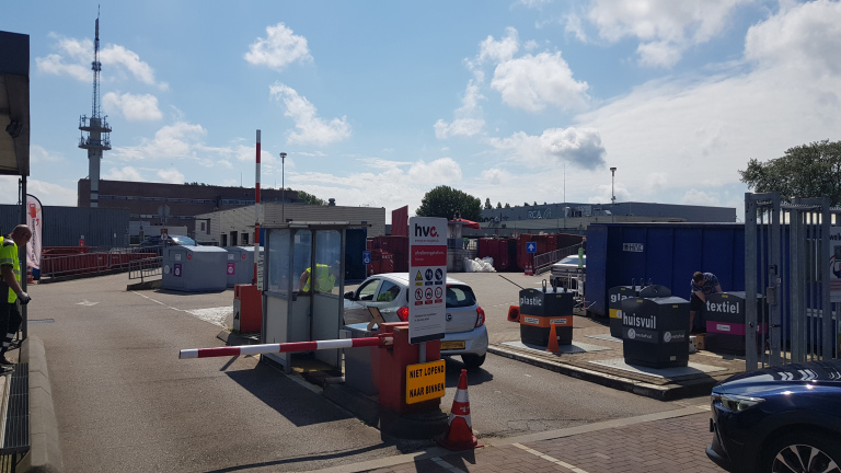 Afvalbrengstation met auto’s, personen en verschillende afvalcontainers, inclusief huishoudelijk afval, plastic, glas en textiel.